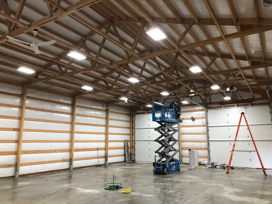 The inside of a pole barn with LED high-bay lights on in the trusses, white liner walls and a scissor lift on the floor
