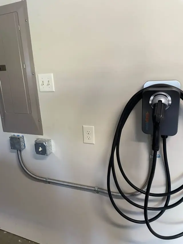 A wall-mounted EV charger with its cable coiled, next to a breaker panel in a garage, with metal conduit running along the wall