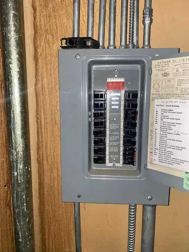 An older gray breaker panel on a wood wall with metal conduit above it, an old panel we have replaced